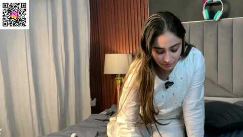 Media: Video of a young woman with long, dark hair in a white cardigan, sitting on a bed with grey sheets. The room features a wooden headboard, white curtains, and a bedside table lamp.