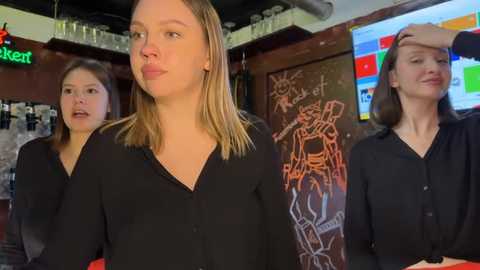 Video of three women in a bar: one blonde in black, one brunette in black, and one with green hair in black, in a dimly lit setting with a colorful mural and bar counter.