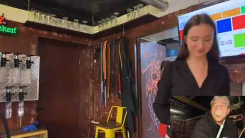 Video of a woman in a black dress with glasses, standing in a cluttered, dimly lit room with a large anatomical chart, shelves of glasses, and a TV playing colorful graphics.