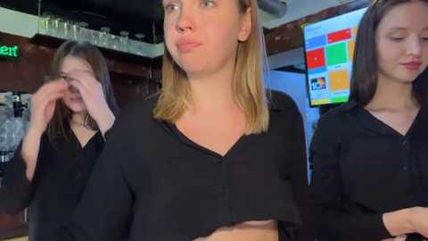 Video of three young women in black shirts, one with a cheeky pose, another hiding eyes, in a modern kitchen with colorful computer screens in the background.