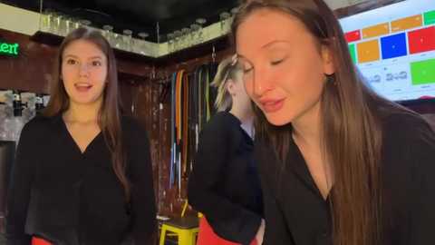Video of two women in black shirts and long brown hair, one smiling, one looking down, in a cluttered bar with colorful signs and shelves.