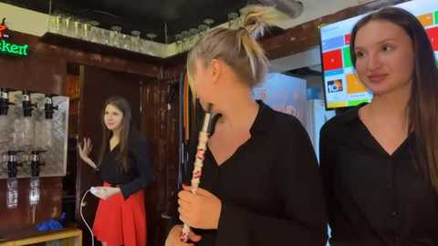 Video of two young women in black shirts and one in a red skirt, standing in a dimly-lit bar with wooden walls, shelves, and a TV screen showing a colorful grid.