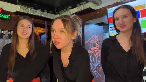 Video of three young Caucasian women in black shirts, one with a ponytail, standing in a bar with a colorful mural, glassware, and a TV.