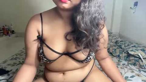 Media: Video of a plus-size South Asian woman with medium skin tone, wearing a leopard print bra, sitting on a bed with floral bedding. Background shows a cluttered room with a wall-mounted light switch.