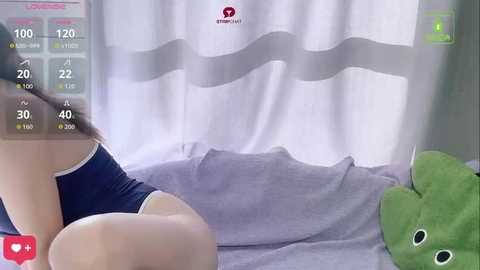 Video of a woman with light skin wearing a blue tank top and shorts, sitting on a bed with a green stuffed animal. A white curtain with a wavy pattern is in the background.