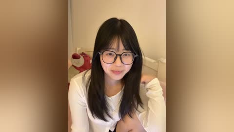 A video of a young Asian woman with straight black hair, wearing large black-framed glasses and a white top, sitting on a bed in a beige room with a stuffed animal.