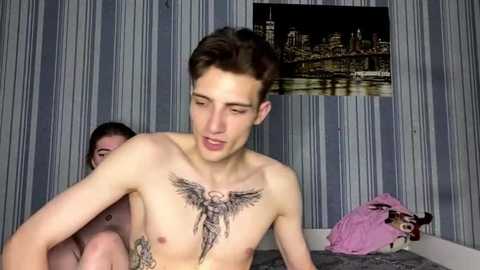 Video of a shirtless young man with short brown hair and a large angel wing tattoo on his chest, in a bedroom with striped wallpaper and a cityscape photo on the wall.