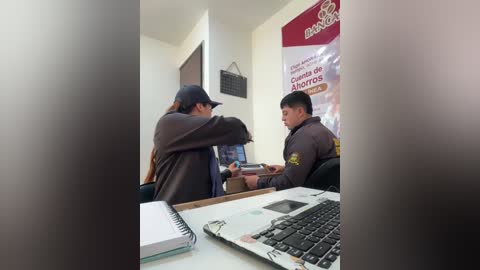 Video of a security guard in a grey uniform, interacting with a woman in a brown jacket and cap, at a desk with a laptop and papers, in a dimly lit office.
