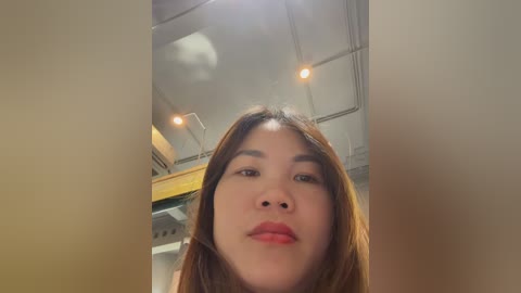 Video of a young Asian woman with straight, shoulder-length brown hair, wearing light makeup and a red lipstick, standing in a modern, dimly lit room with exposed ceiling beams and recessed lights.