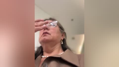 Video of a middle-aged woman with glasses, wearing a beige jacket and pearl necklace, shielding her eyes from the bright light.