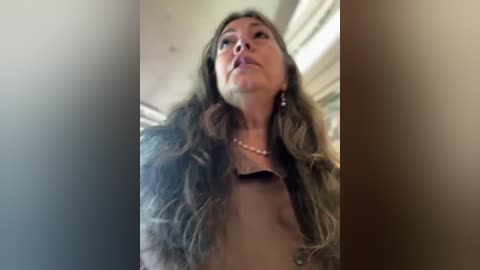 Video of a middle-aged woman with long, wavy brown hair, wearing a beige top and a pearl necklace, looking upward with a serious expression.