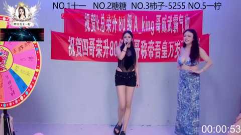 Video of three women on stage, one in a black bodysuit, another in a blue dress, and a third in a colorful dress. Red Chinese banners, vibrant background, and a colorful wheel of fortune.