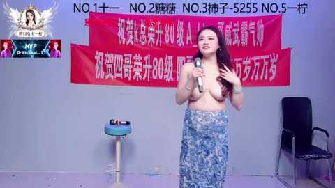 Video of an Asian woman with long hair, topless, holding a microphone, speaking against a backdrop with red Chinese text. Background includes a chair and a podium with a box.