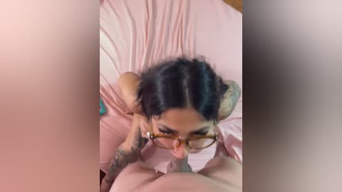 A video of a woman with dark hair, wearing glasses, performing oral sex on a man. Both are on a bed with pink sheets, and the man has a tattooed arm visible.