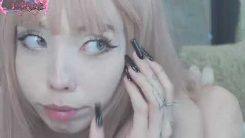 A close-up video of a young woman with light skin, pink hair, and dramatic black winged eyeliner, speaking on a phone. Background is blurred with a hint of a textured wall.