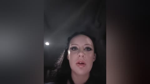 Video of a pale woman with long black hair, wearing dark makeup, looking directly at the camera. The background is dimly lit with a single bright light source on the left, creating a dramatic effect.