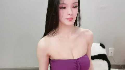 Video of a young East Asian woman with long black hair, fair skin, and a slender physique, wearing a strapless purple dress. Background shows a gray wall and a white fur rug.