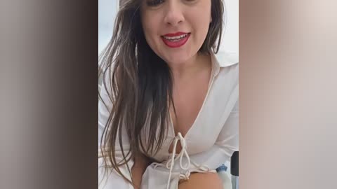 Video of a smiling woman with long brown hair, wearing a white blouse, leaning forward with a playful expression.