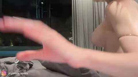 A video of a woman with fair skin, topless, showcasing her medium-sized breasts, while a hand, possibly hers, is gesturing in the foreground. The background features a dimly lit room with vertical blinds.