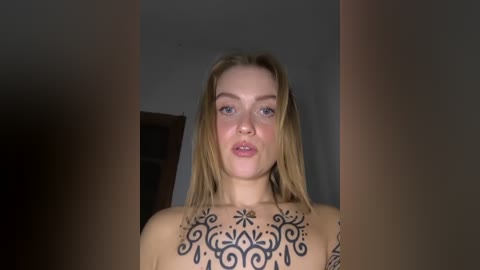 Video of a topless, light-skinned woman with long, blonde hair, wearing a black, intricate, floral tattoo on her chest. She has blue eyes and is standing indoors, partially obscured by blurry, brownish objects.