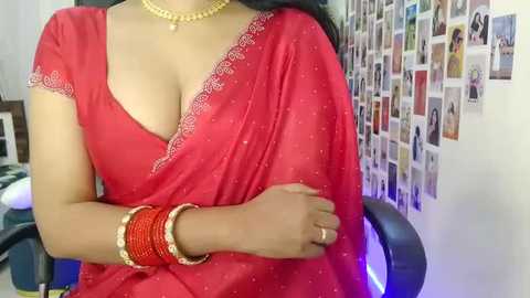 Video of a woman with medium brown skin, wearing a red saree with gold embroidery, gold bangles, and a necklace, seated in a chair, surrounded by a collage of photos on a white wall.