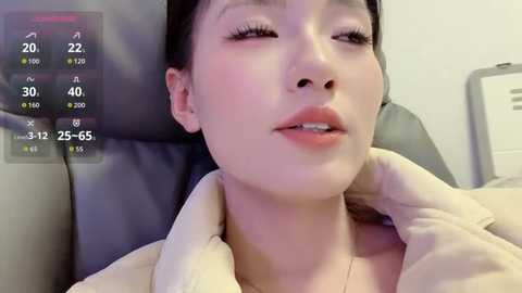 A video of an Asian woman with fair skin, dark hair, and closed eyes, wearing a beige jacket, lying on a grey leather chair, with a digital screen showing temperature and time.