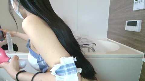A video shows a woman with long, straight black hair, wearing a plaid apron, using a bottle to dye her hair in a modern bathroom with a bathtub, white tiles, and a wall-mounted heater.