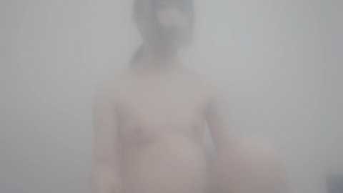 Blurry video of a nude, pale-skinned person with long hair, standing in a dimly lit, misty room. The image is soft and indistinct, with no discernible features or background details.