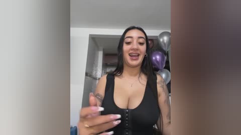 Media: Video of a Latina woman with long black hair, wearing a black top, holding a phone, surrounded by balloons in a brightly lit room.