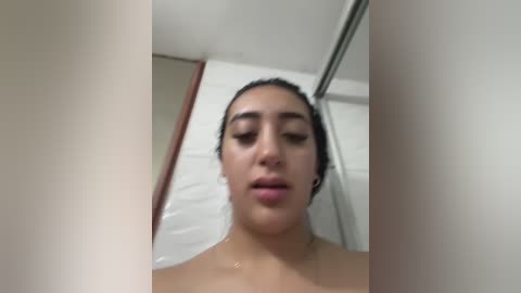 Video of a Latina woman with wet hair, dark eyes, and full lips, standing in a bathroom. She has medium skin tone and is topless, with the focus on her face and upper body.
