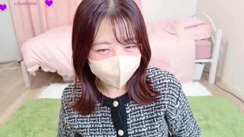 Video of an Asian woman with long, straight, dark hair, wearing a beige face mask, a black-and-white patterned coat, and seated on a green rug in a pastel bedroom.