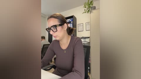 Video of a woman with glasses, brown hair tied back, wearing a long-sleeved maroon top, seated at a desk in an office, surrounded by bookshelves and potted plants.