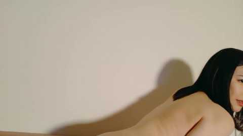 Media: Video of a nude woman with pale skin and long black hair, lying face down on a bed, against a plain beige wall.