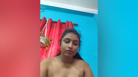 A video of a young South Asian woman with medium-brown skin, long dark hair, and full lips, standing nude in a brightly lit room with blue and red walls.