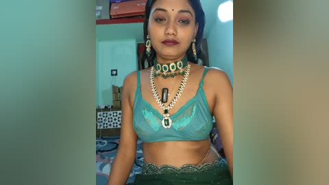Video of a South Asian woman with medium brown skin, wearing a turquoise bra, green skirt, and traditional jewelry, including a choker and large earrings, in a modestly furnished room.
