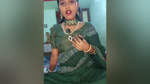 Video of a South Asian woman in a green sari with intricate silver embroidery, wearing heavy gold jewelry, including a necklace with large pendant, and earrings, sitting on a bed in a modestly furnished room.