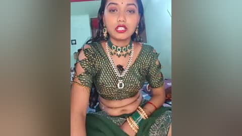 Video of an Indian woman in traditional green saree with gold embroidery, adorned with matching green jewelry and bindi, seated in a simple indoor setting.