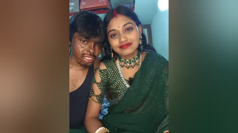Video of a woman with a green saree and matching jewelry, standing beside a man with a bandaged face, both smiling, in a simple room with a red cabinet in the background.