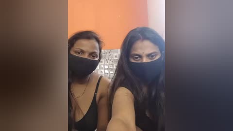 Video of two women with long dark hair, wearing black face masks, black tank tops, and gold necklaces, standing side by side in a brightly lit bathroom with orange and white tiled walls.