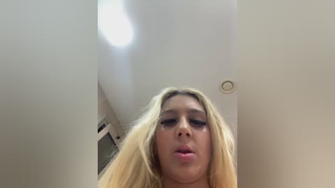 Video of a young, light-skinned woman with long, straight blonde hair and heavy makeup, standing in a dimly lit room with a white ceiling and a small light fixture.
