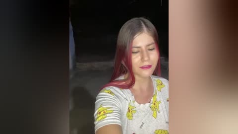 Video of a young woman with long, straight, dyed red hair, wearing a white t-shirt with yellow cartoon characters, smiling, standing in a dark, blurry background.