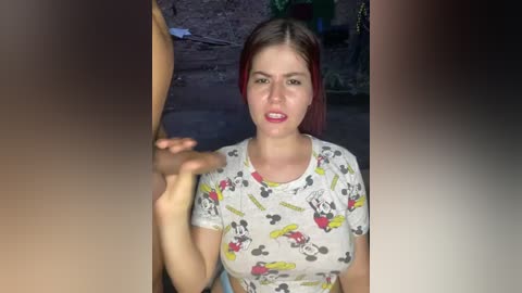 A video of a young woman with fair skin and red hair, wearing a Minnie Mouse T-shirt, holding a hand in front of her face, standing in a dimly lit, outdoor setting.