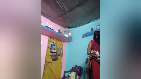 A video of a small, brightly lit room with blue walls and a yellow door. A woman in a red sari stands near a blue shelf, holding a white object.