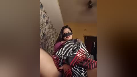 Media: Video of a woman in a black-and-white striped dress, wearing a black blindfold, sitting on a couch. Background features a patterned wall and a door. The scene suggests a playful, intimate setting.