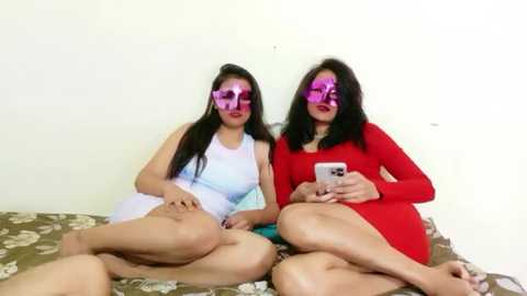 Video of two women with light skin, long black hair, wearing pink masks, sitting on a floral-patterned bed, one in a white dress, the other in a red top, holding a phone.