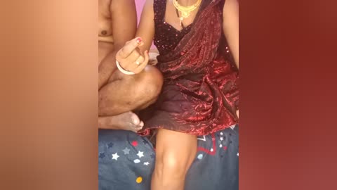 Video of a naked man with light brown skin, lifting a woman with medium brown skin in a red sari, on a blue bed with star patterns.