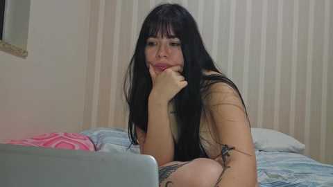 Media: Video of a young woman with long black hair and fair skin, lying on a bed, looking sad, wearing a black lace bra, in a bedroom with beige curtains.