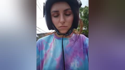 Video of a young woman with light skin, wearing a tie-dye shirt, large black headphones, and a neutral expression. She's in an outdoor setting with blurred greenery and a building in the background.