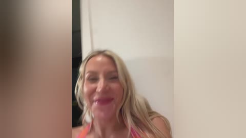 Video of a blonde woman with fair skin and long hair, wearing a pink tank top, laughing, blurred in the background.