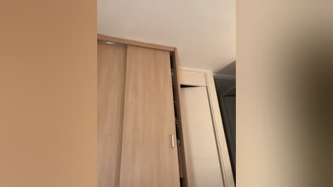 Video of a minimalist, beige-walled bedroom featuring a sliding wooden wardrobe with a sleek, silver handle. The wardrobe is partially open, revealing a white interior. The room has a soft, neutral ambiance.